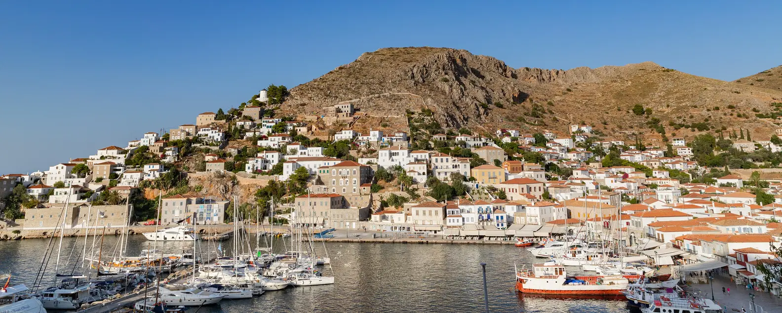 Le port d’Hydra avec des voiliers et des demeures en pierre qui montent la pente sous une montagne nue