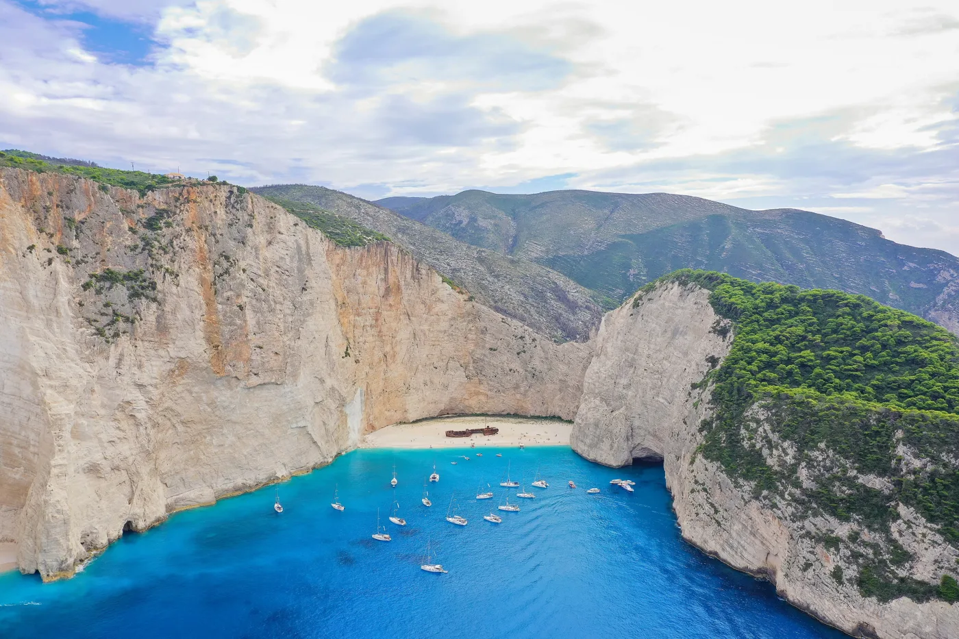 Navagio from above, with the cliffs, the rusty ship on the sand and boats in the blue water