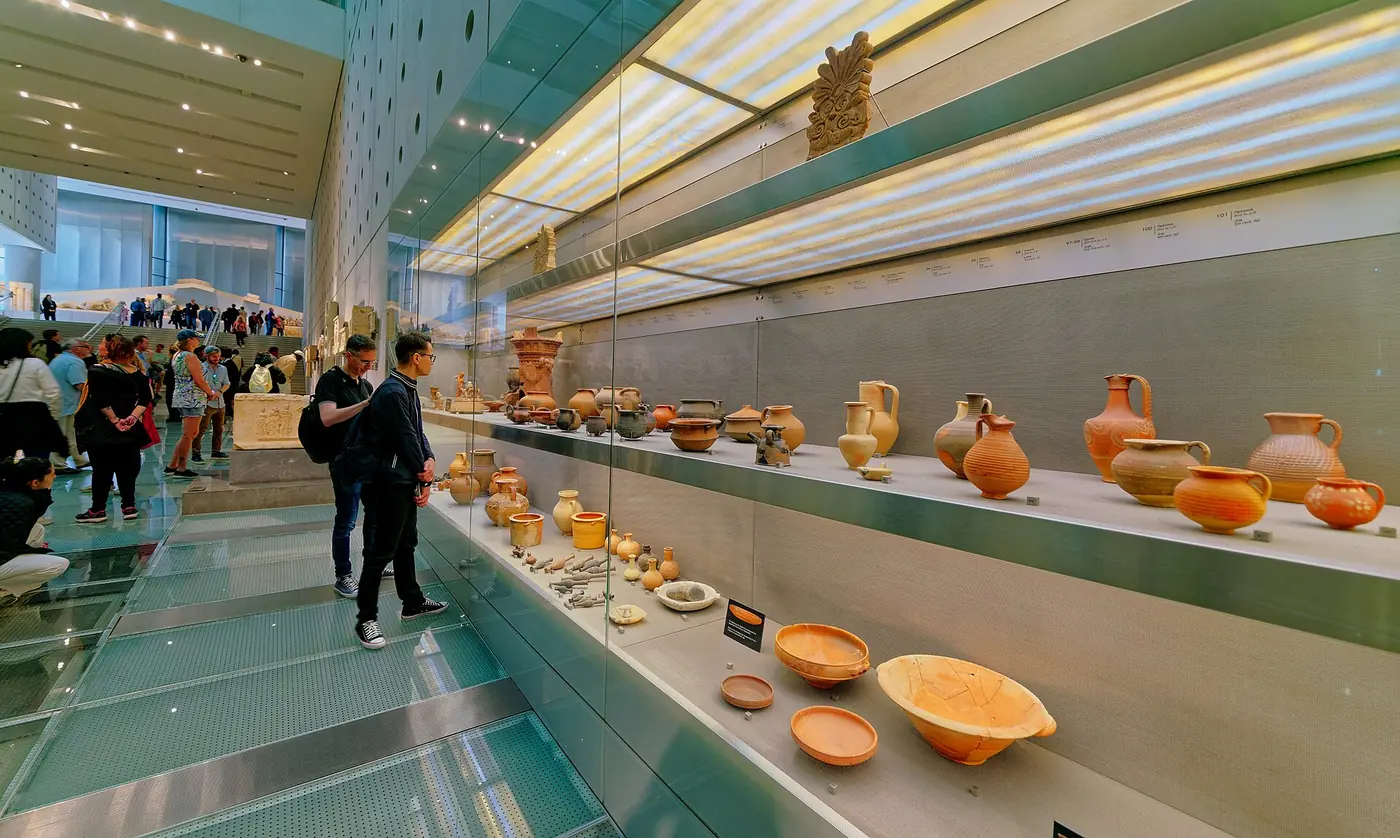 参观者走在玻璃地面上，旁边是展出卫城圣所陶器的展柜