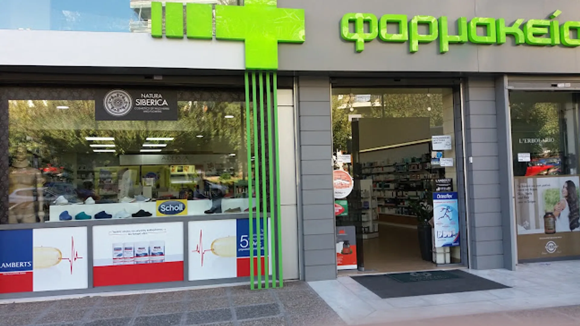 An illuminated green-cross pharmacy sign on a quiet Athenian street at dusk